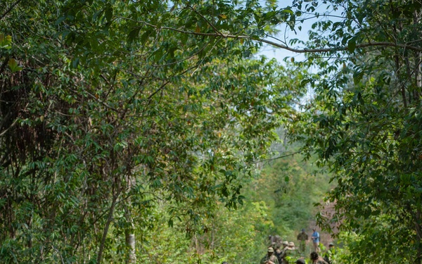A mixed group of military personnel conducting a patrol along a narrow dirt trail through dense tropical jungle vegetation. The group includes soldiers in at least two distinct camouflage patterns, suggesting a multinational joint training exercise or operation. The central figure wears tan/multicam uniform with a boonie hat and carries a rifle with tactical accessories.