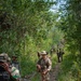 A mixed group of military personnel conducting a patrol along a narrow dirt trail through dense tropical jungle vegetation. The group includes soldiers in at least two distinct camouflage patterns, suggesting a multinational joint training exercise or operation. The central figure wears tan/multicam uniform with a boonie hat and carries a rifle with tactical accessories.