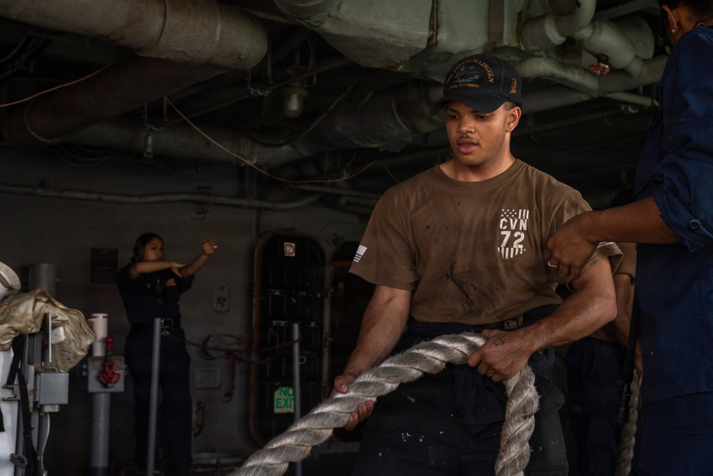 A U.S. Navy sailor aboard USS Abraham Lincoln (CVN-72) is hauling a large mooring line in what appears to be a hangar bay or below-deck working area. The sailor is wearing a CVN-72 shirt and a ship's ball cap, with at least two other crew members visible in the background. The scene depicts routine line-handling or mooring operations aboard the aircraft carrier.