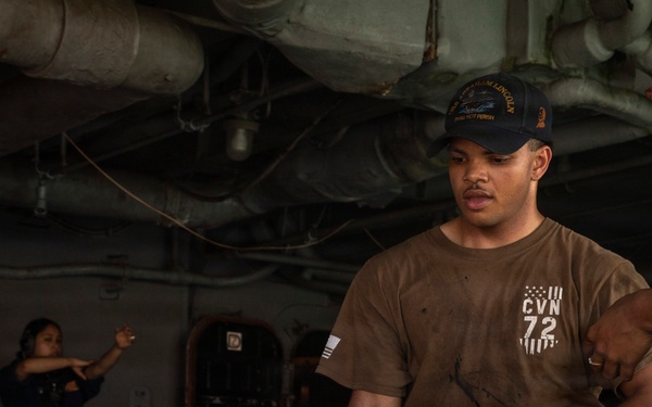 A U.S. Navy sailor aboard USS Abraham Lincoln (CVN-72) is hauling a large mooring line in what appears to be a hangar bay or below-deck working area. The sailor is wearing a CVN-72 shirt and a ship's ball cap, with at least two other crew members visible in the background. The scene depicts routine line-handling or mooring operations aboard the aircraft carrier.