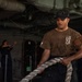 A U.S. Navy sailor aboard USS Abraham Lincoln (CVN-72) is hauling a large mooring line in what appears to be a hangar bay or below-deck working area. The sailor is wearing a CVN-72 shirt and a ship's ball cap, with at least two other crew members visible in the background. The scene depicts routine line-handling or mooring operations aboard the aircraft carrier.