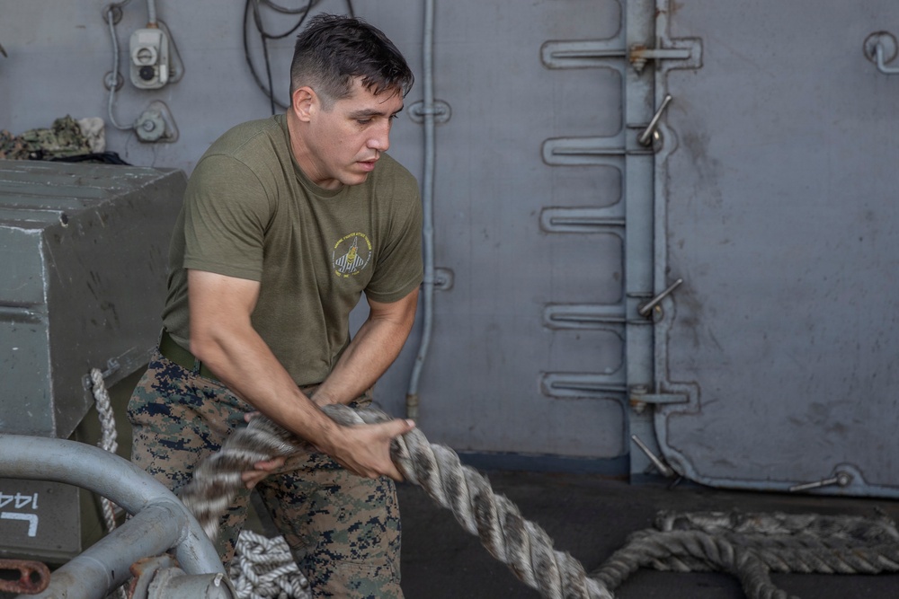 A male service member wearing a U.S. Marine Corps olive drab t-shirt and MARPAT digital camouflage trousers is handling a large mooring or utility rope aboard what appears to be a naval vessel. He is gripping the thick rope with both hands in a focused, physical effort. The setting is an exterior deck area of a ship with gray metal walls, pipes, and fittings visible in the background.