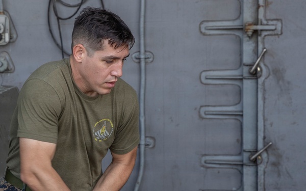 A male service member wearing a U.S. Marine Corps olive drab t-shirt and MARPAT digital camouflage trousers is handling a large mooring or utility rope aboard what appears to be a naval vessel. He is gripping the thick rope with both hands in a focused, physical effort. The setting is an exterior deck area of a ship with gray metal walls, pipes, and fittings visible in the background.