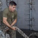 A male service member wearing a U.S. Marine Corps olive drab t-shirt and MARPAT digital camouflage trousers is handling a large mooring or utility rope aboard what appears to be a naval vessel. He is gripping the thick rope with both hands in a focused, physical effort. The setting is an exterior deck area of a ship with gray metal walls, pipes, and fittings visible in the background.