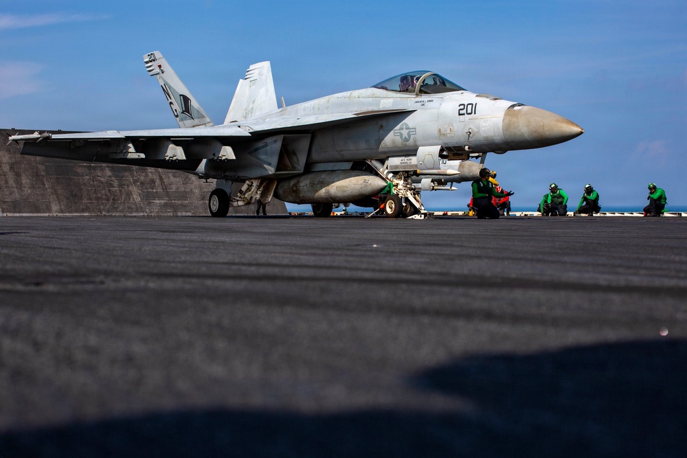 A U.S. Navy F/A-18 Super Hornet (Bureau Number 201) is positioned on an aircraft carrier flight deck, apparently being prepared for launch. Several deck crew members in green jerseys and helmets are crouched near the aircraft in standard pre-launch positions. The image is taken from a low deck-level angle, emphasizing the scale of the aircraft and the carrier's flight deck.