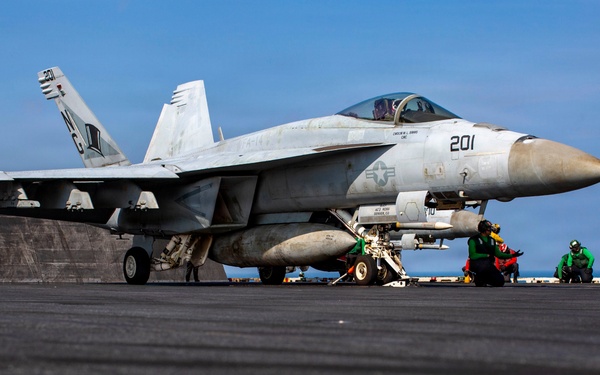 A U.S. Navy F/A-18 Super Hornet (Bureau Number 201) is positioned on an aircraft carrier flight deck, apparently being prepared for launch. Several deck crew members in green jerseys and helmets are crouched near the aircraft in standard pre-launch positions. The image is taken from a low deck-level angle, emphasizing the scale of the aircraft and the carrier's flight deck.