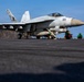 A U.S. Navy F/A-18 Super Hornet (Bureau Number 201) is positioned on an aircraft carrier flight deck, apparently being prepared for launch. Several deck crew members in green jerseys and helmets are crouched near the aircraft in standard pre-launch positions. The image is taken from a low deck-level angle, emphasizing the scale of the aircraft and the carrier's flight deck.