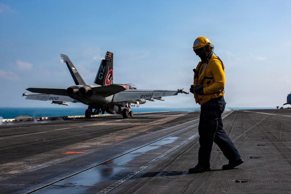 A U.S. Navy flight deck crew member in yellow jersey and helmet stands on an aircraft carrier flight deck as an F/A-18 Super Hornet prepares for or completes a catapult launch. The aircraft bears the tail code 'NG' and modex '200', indicating it belongs to a carrier air wing squadron. The scene takes place at sea under clear skies with the ocean visible in the background.