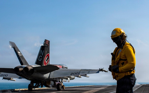 A U.S. Navy flight deck crew member in yellow jersey and helmet stands on an aircraft carrier flight deck as an F/A-18 Super Hornet prepares for or completes a catapult launch. The aircraft bears the tail code 'NG' and modex '200', indicating it belongs to a carrier air wing squadron. The scene takes place at sea under clear skies with the ocean visible in the background.