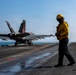 A U.S. Navy flight deck crew member in yellow jersey and helmet stands on an aircraft carrier flight deck as an F/A-18 Super Hornet prepares for or completes a catapult launch. The aircraft bears the tail code 'NG' and modex '200', indicating it belongs to a carrier air wing squadron. The scene takes place at sea under clear skies with the ocean visible in the background.