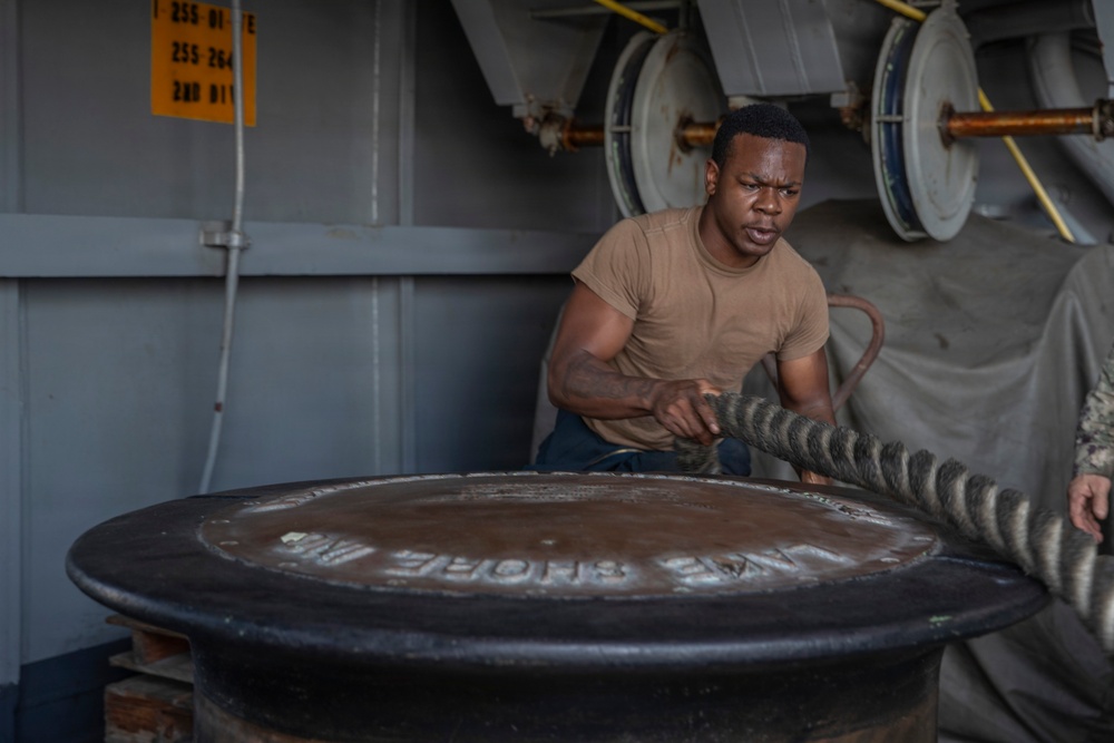 A sailor or military service member in a tan/brown t-shirt is working with a large mooring line on what appears to be a naval vessel. He is exerting significant physical effort to handle the thick rope around a large bollard or capstan. The setting is clearly a ship's deck with naval equipment visible in the background.