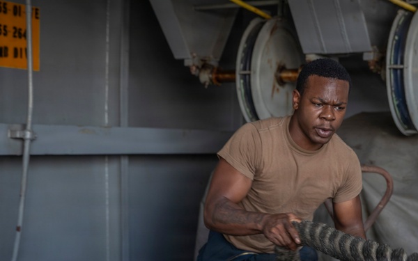 A sailor or military service member in a tan/brown t-shirt is working with a large mooring line on what appears to be a naval vessel. He is exerting significant physical effort to handle the thick rope around a large bollard or capstan. The setting is clearly a ship's deck with naval equipment visible in the background.