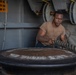A sailor or military service member in a tan/brown t-shirt is working with a large mooring line on what appears to be a naval vessel. He is exerting significant physical effort to handle the thick rope around a large bollard or capstan. The setting is clearly a ship's deck with naval equipment visible in the background.