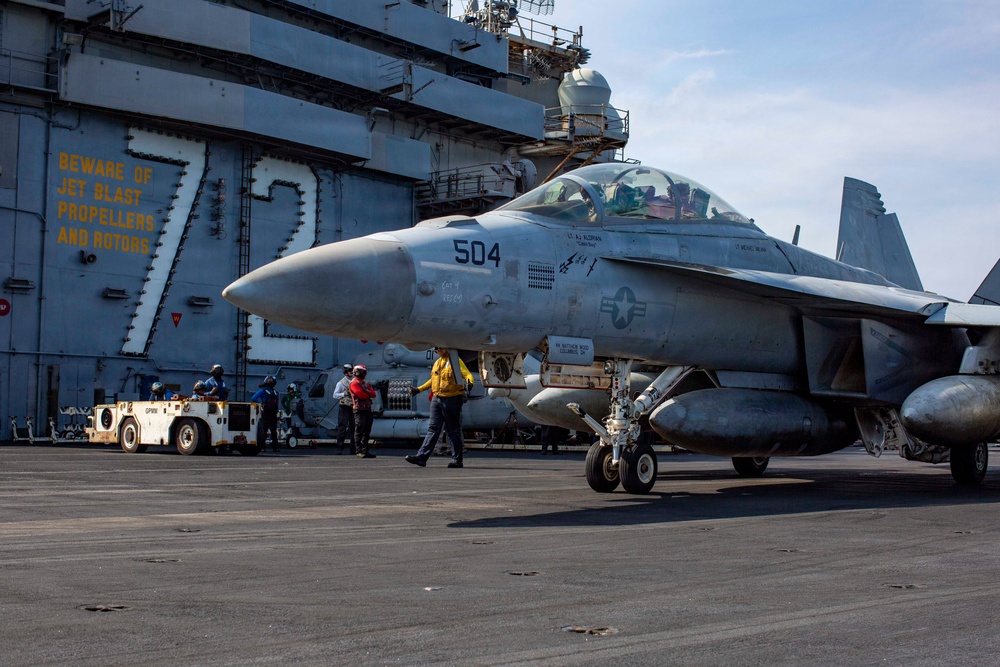 A U.S. Navy F/A-18 Super Hornet (bureau number 504) is positioned on the flight deck of an aircraft carrier, surrounded by deck crew in color-coded jerseys. The carrier's island structure is visible in the background with a large yellow '7' and safety warning text reading 'BEWARE OF JET BLAST PROPELLERS AND ROTORS.' Deck personnel are actively conducting pre-flight or post-flight operations around the aircraft.