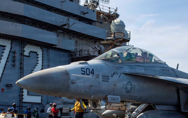 A U.S. Navy F/A-18 Super Hornet (bureau number 504) is positioned on the flight deck of an aircraft carrier, surrounded by deck crew in color-coded jerseys. The carrier's island structure is visible in the background with a large yellow '7' and safety warning text reading 'BEWARE OF JET BLAST PROPELLERS AND ROTORS.' Deck personnel are actively conducting pre-flight or post-flight operations around the aircraft.