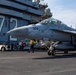 A U.S. Navy F/A-18 Super Hornet (bureau number 504) is positioned on the flight deck of an aircraft carrier, surrounded by deck crew in color-coded jerseys. The carrier's island structure is visible in the background with a large yellow '7' and safety warning text reading 'BEWARE OF JET BLAST PROPELLERS AND ROTORS.' Deck personnel are actively conducting pre-flight or post-flight operations around the aircraft.