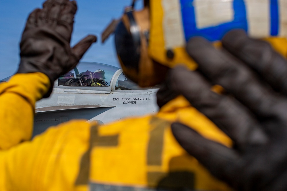 A naval aviation deck crew member in yellow flight deck gear and black gloves signals to a pilot seated in a military fighter jet cockpit. The aircraft fuselage displays the name 'ENS JESSE GRAVLEY' and 'GUNNER', indicating the assigned pilot and crew designation. The scene appears to be on an aircraft carrier flight deck during pre-launch or taxi operations.