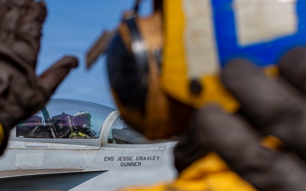 A naval aviation deck crew member in yellow flight deck gear and black gloves signals to a pilot seated in a military fighter jet cockpit. The aircraft fuselage displays the name 'ENS JESSE GRAVLEY' and 'GUNNER', indicating the assigned pilot and crew designation. The scene appears to be on an aircraft carrier flight deck during pre-launch or taxi operations.