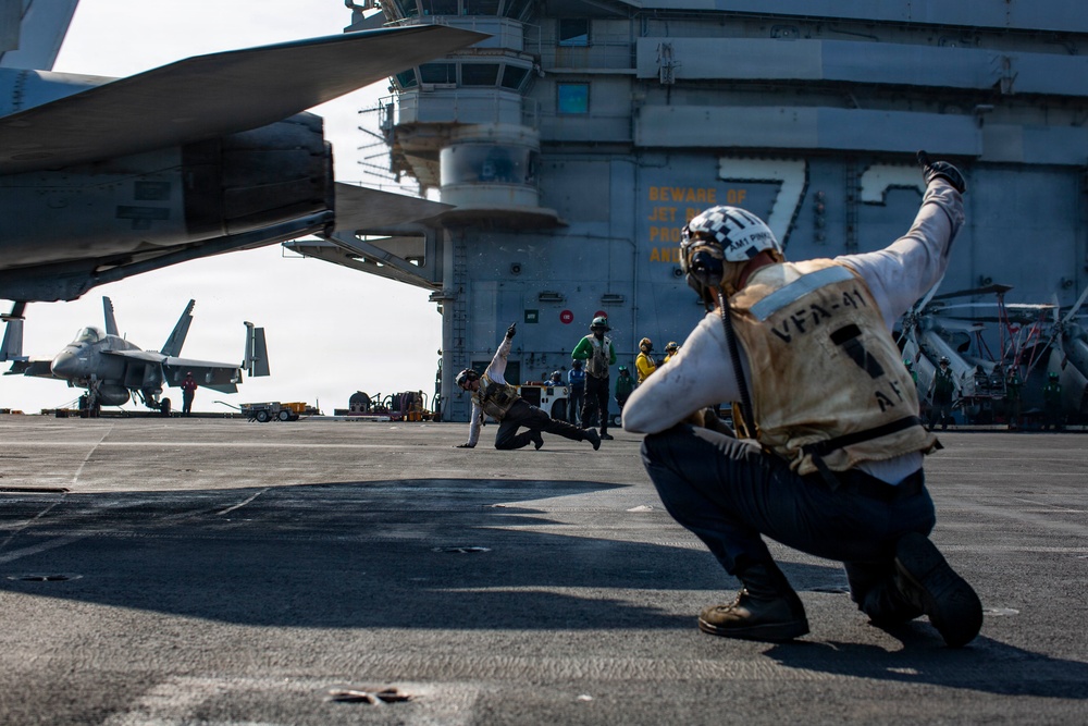 The image shows flight deck operations aboard a U.S. Navy aircraft carrier, with crew members performing catapult launch signaling procedures. A shooter or deck crew member in the foreground is crouching in the characteristic launch signal pose, wearing a vest marked 'VFA-41' with a checkered helmet. Multiple F/A-18 aircraft are visible on the flight deck in the background.