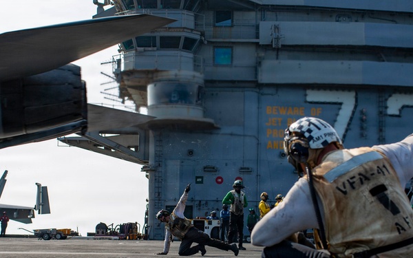 The image shows flight deck operations aboard a U.S. Navy aircraft carrier, with crew members performing catapult launch signaling procedures. A shooter or deck crew member in the foreground is crouching in the characteristic launch signal pose, wearing a vest marked 'VFA-41' with a checkered helmet. Multiple F/A-18 aircraft are visible on the flight deck in the background.