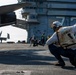 The image shows flight deck operations aboard a U.S. Navy aircraft carrier, with crew members performing catapult launch signaling procedures. A shooter or deck crew member in the foreground is crouching in the characteristic launch signal pose, wearing a vest marked 'VFA-41' with a checkered helmet. Multiple F/A-18 aircraft are visible on the flight deck in the background.
