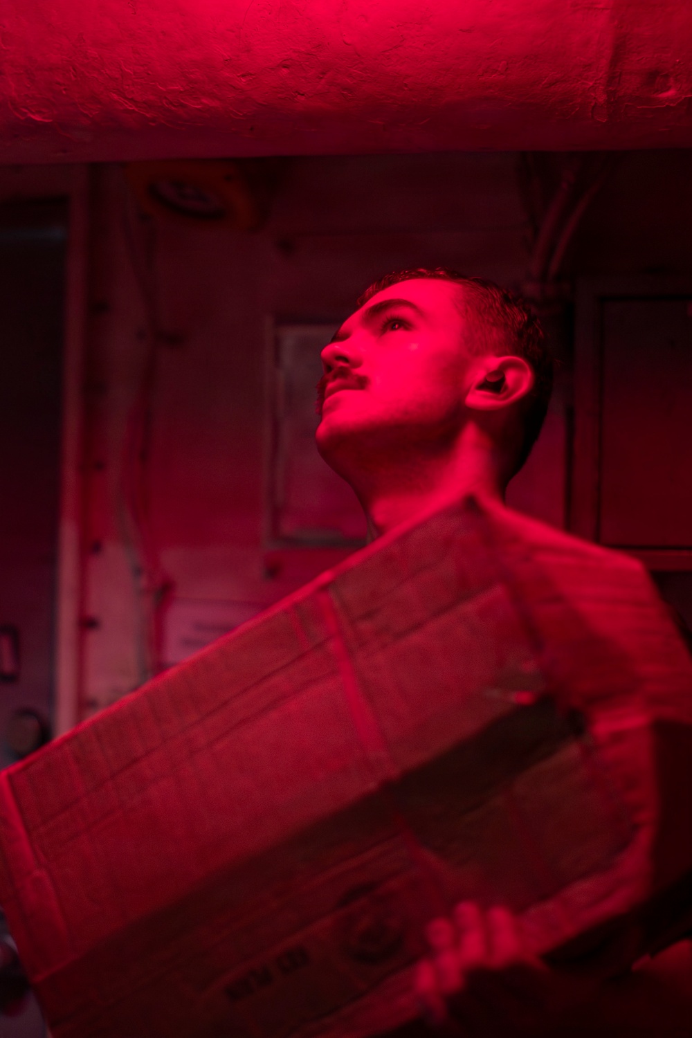 A young man is photographed under intense red lighting in what appears to be a confined, industrial or military interior space. He is carrying a cardboard box and looking upward, suggesting he is navigating a storage or cargo area. The dominant red illumination creates a dramatic, low-visibility atmosphere consistent with night-operations lighting found on naval vessels or similar facilities.