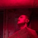 A young man is photographed under intense red lighting in what appears to be a confined, industrial or military interior space. He is carrying a cardboard box and looking upward, suggesting he is navigating a storage or cargo area. The dominant red illumination creates a dramatic, low-visibility atmosphere consistent with night-operations lighting found on naval vessels or similar facilities.