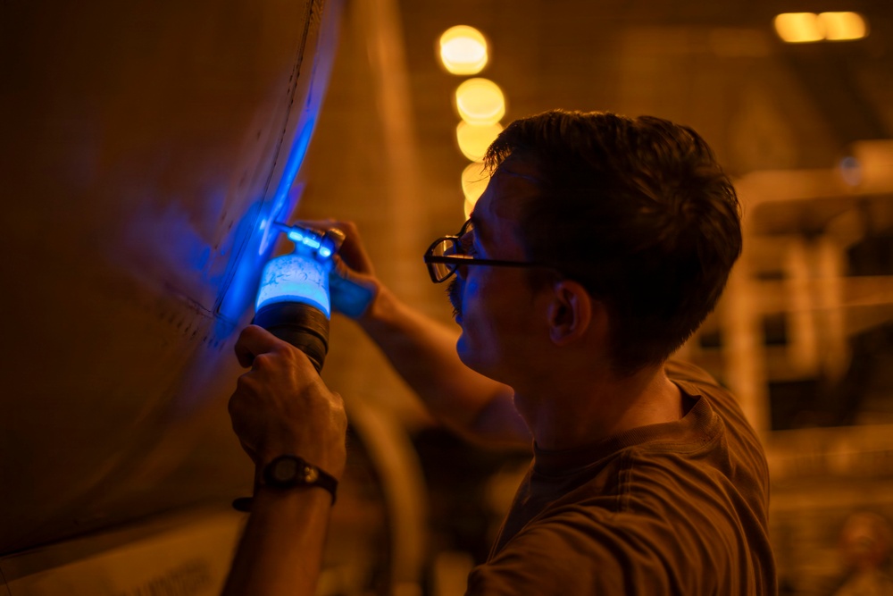 A man wearing glasses and a wristwatch is using a blue UV/ultraviolet flashlight to inspect a large curved surface, likely an aircraft fuselage or industrial equipment. The inspection appears to be conducted in a low-light environment illuminated by warm orange ambient lighting in the background. The blue UV light suggests a non-destructive testing or maintenance inspection procedure.