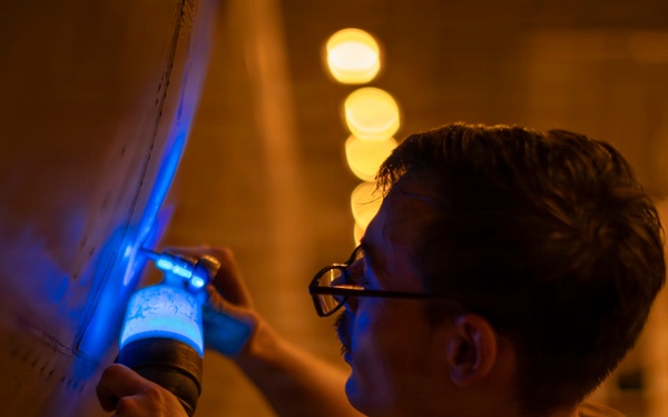 A man wearing glasses and a wristwatch is using a blue UV/ultraviolet flashlight to inspect a large curved surface, likely an aircraft fuselage or industrial equipment. The inspection appears to be conducted in a low-light environment illuminated by warm orange ambient lighting in the background. The blue UV light suggests a non-destructive testing or maintenance inspection procedure.