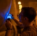 A man wearing glasses and a wristwatch is using a blue UV/ultraviolet flashlight to inspect a large curved surface, likely an aircraft fuselage or industrial equipment. The inspection appears to be conducted in a low-light environment illuminated by warm orange ambient lighting in the background. The blue UV light suggests a non-destructive testing or maintenance inspection procedure.
