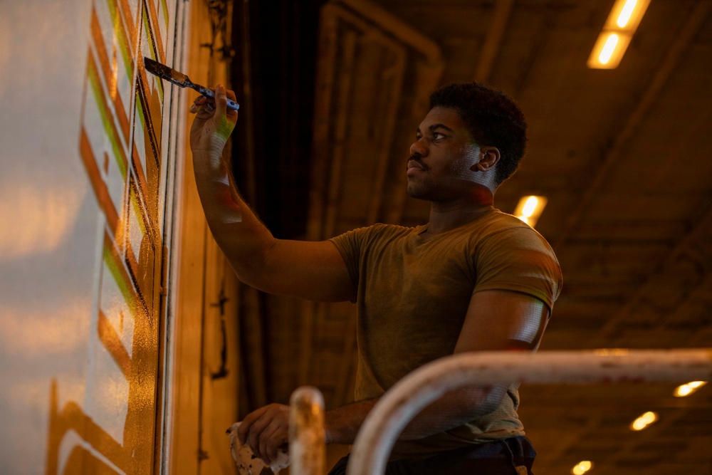 A young man is painting a mural or decorative design on a large wall surface in what appears to be an industrial or warehouse-type space. He is using a paintbrush with one hand while holding a cloth or rag in the other, applying orange, white, and green geometric patterns. The scene is lit with warm amber overhead lighting, creating a dramatic, low-light atmosphere.