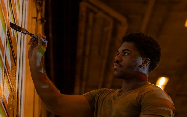 A young man is painting a mural or decorative design on a large wall surface in what appears to be an industrial or warehouse-type space. He is using a paintbrush with one hand while holding a cloth or rag in the other, applying orange, white, and green geometric patterns. The scene is lit with warm amber overhead lighting, creating a dramatic, low-light atmosphere.