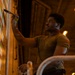 A young man is painting a mural or decorative design on a large wall surface in what appears to be an industrial or warehouse-type space. He is using a paintbrush with one hand while holding a cloth or rag in the other, applying orange, white, and green geometric patterns. The scene is lit with warm amber overhead lighting, creating a dramatic, low-light atmosphere.