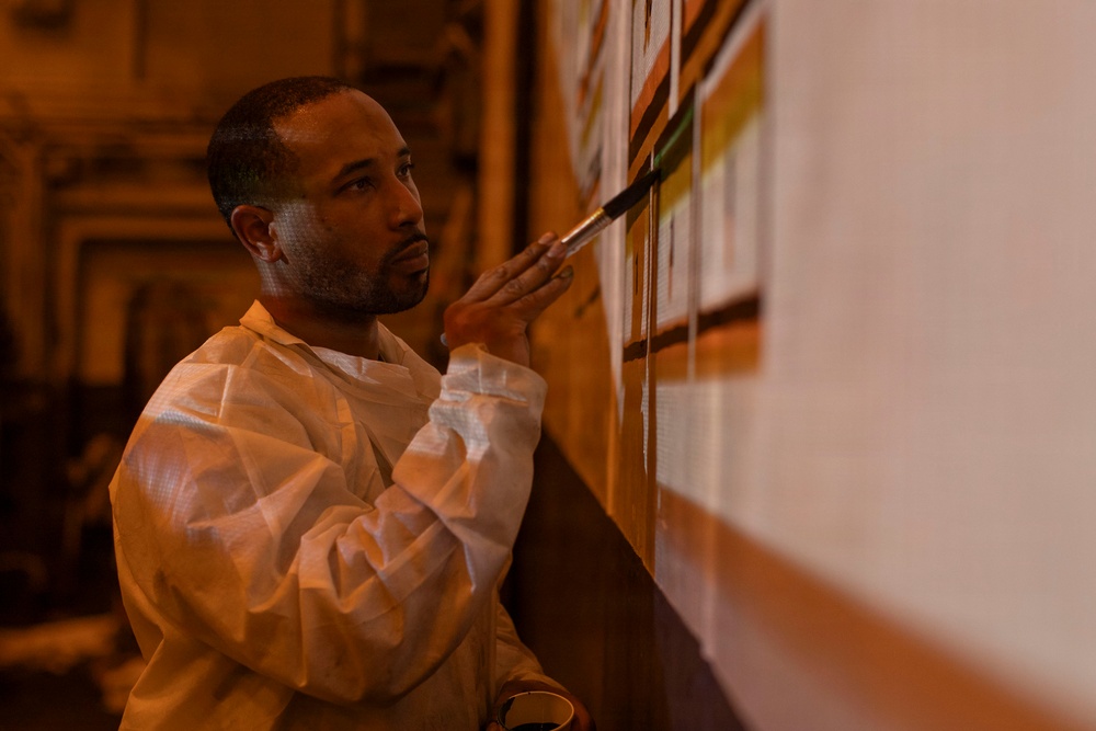 A man wearing a white protective coverall suit is carefully applying paint to a wall using a fine-tipped brush. He appears to be working on a mural or decorative wall painting, holding what appears to be a small container in his other hand. The scene is lit with warm amber/orange tones, suggesting an interior architectural space.