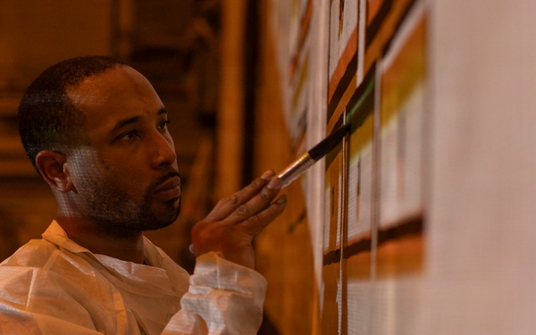 A man wearing a white protective coverall suit is carefully applying paint to a wall using a fine-tipped brush. He appears to be working on a mural or decorative wall painting, holding what appears to be a small container in his other hand. The scene is lit with warm amber/orange tones, suggesting an interior architectural space.