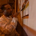 A man wearing a white protective coverall suit is carefully applying paint to a wall using a fine-tipped brush. He appears to be working on a mural or decorative wall painting, holding what appears to be a small container in his other hand. The scene is lit with warm amber/orange tones, suggesting an interior architectural space.