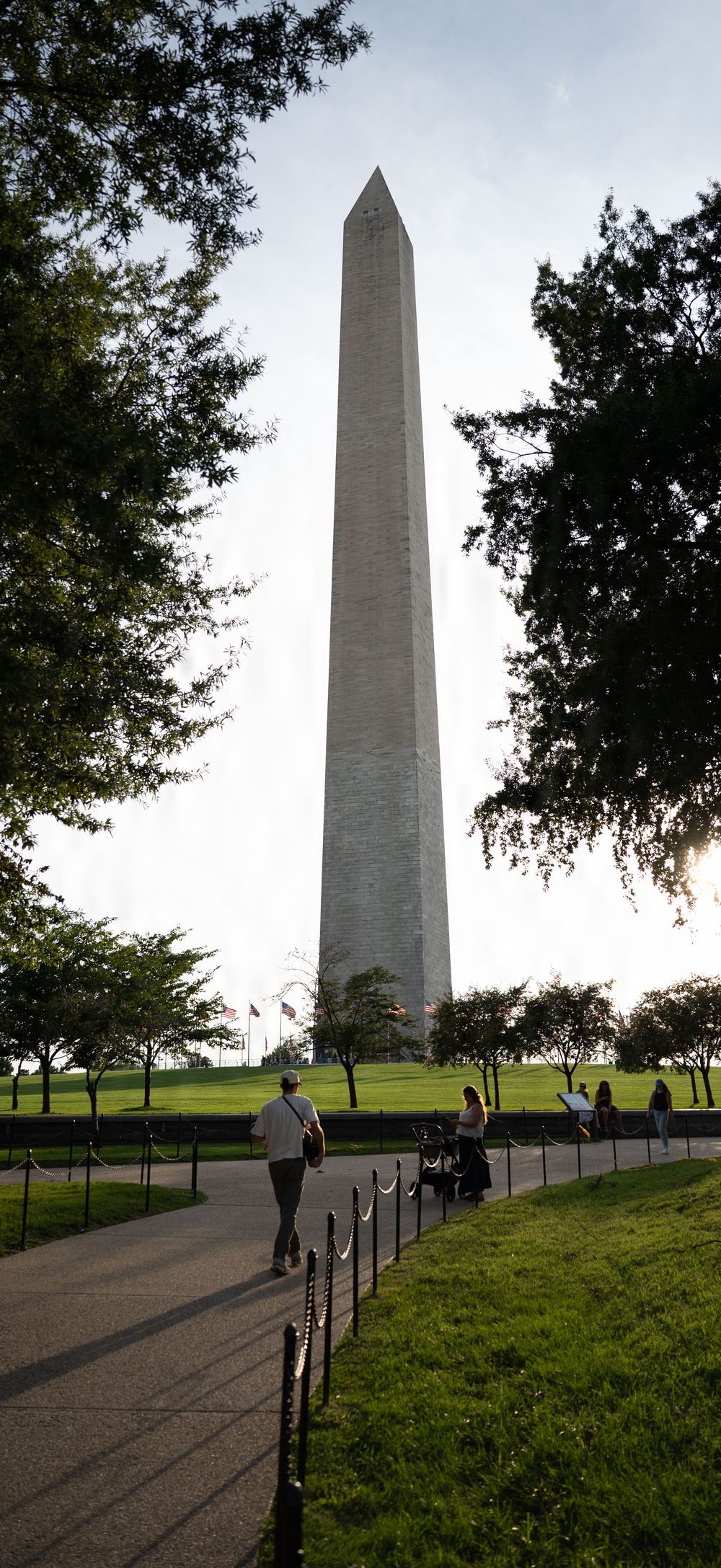 A ground-level view of the Washington Monument in Washington, D.C., photographed from a paved walkway leading toward the base of the obelisk. The image is taken during golden hour with backlit lighting, creating a dramatic silhouette effect against a bright sky. Several visitors are visible on and near the path, with lush green lawns and trees framing the monument on both sides.