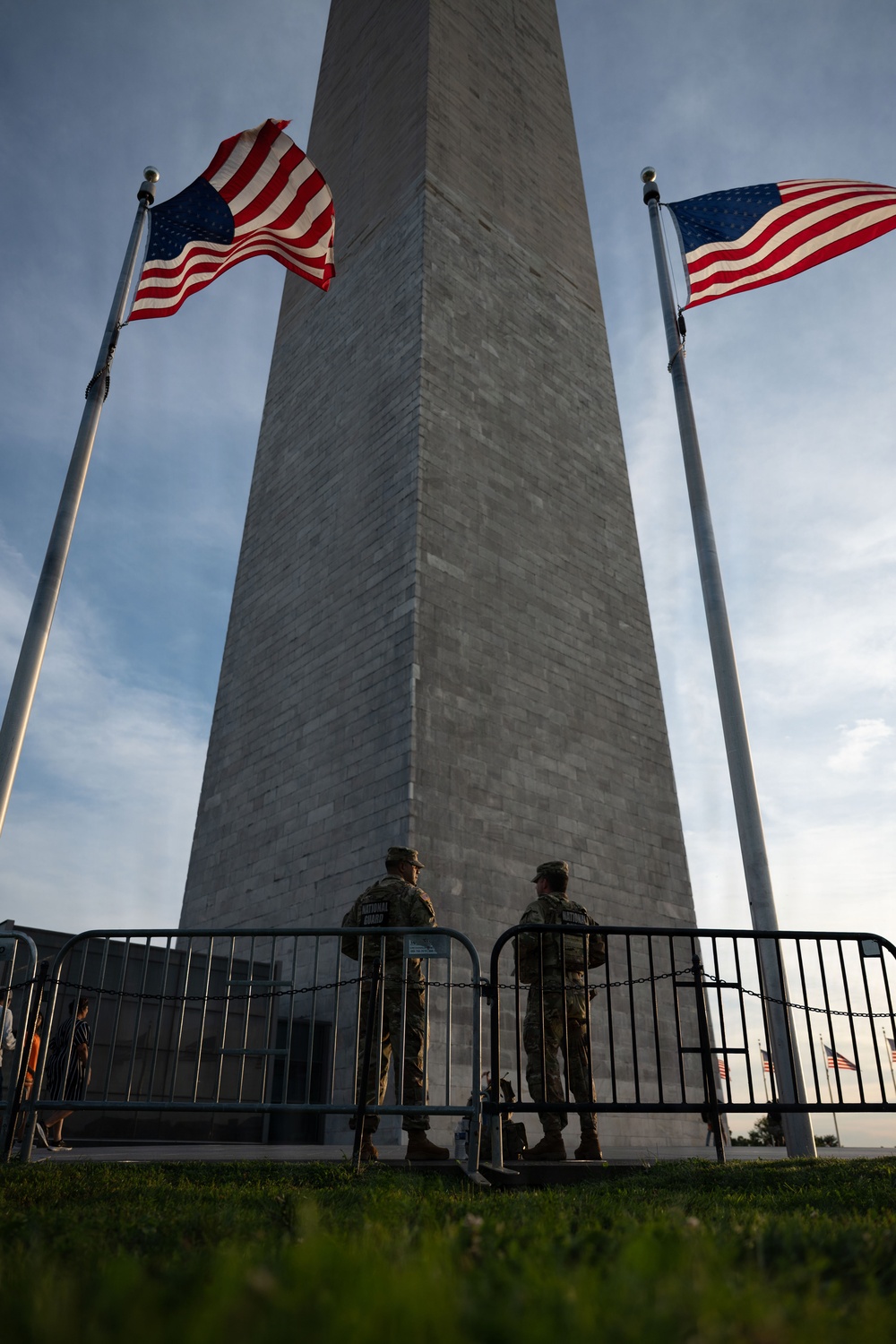 Two National Guard soldiers stand at the base of the Washington Monument in Washington, D.C., behind metal security barriers. Two American flags flank the obelisk on either side, flying in the wind against a partly cloudy sky. The low-angle shot emphasizes the monument's height and the security presence at its entrance.