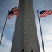 Two National Guard soldiers stand at the base of the Washington Monument in Washington, D.C., behind metal security barriers. Two American flags flank the obelisk on either side, flying in the wind against a partly cloudy sky. The low-angle shot emphasizes the monument's height and the security presence at its entrance.