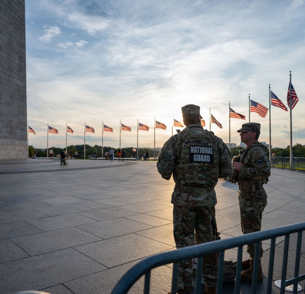 Two National Guard soldiers in camouflage uniforms stand at the base of the Washington Monument on the National Mall in Washington, D.C. They are positioned behind crowd control barriers while a row of American flags flies in the background. The image appears to be taken during evening hours with a partially cloudy sky and warm backlighting.