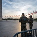 Two National Guard soldiers in camouflage uniforms stand at the base of the Washington Monument on the National Mall in Washington, D.C. They are positioned behind crowd control barriers while a row of American flags flies in the background. The image appears to be taken during evening hours with a partially cloudy sky and warm backlighting.