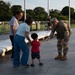 A U.S. Army soldier in full camouflage uniform is bending down to interact with a young toddler in a red shirt, while two adults (likely grandparents) stand nearby watching. The scene takes place on a paved plaza area, possibly a memorial or public monument, during golden hour lighting. The interaction appears warm and friendly, with the soldier extending something toward the child.