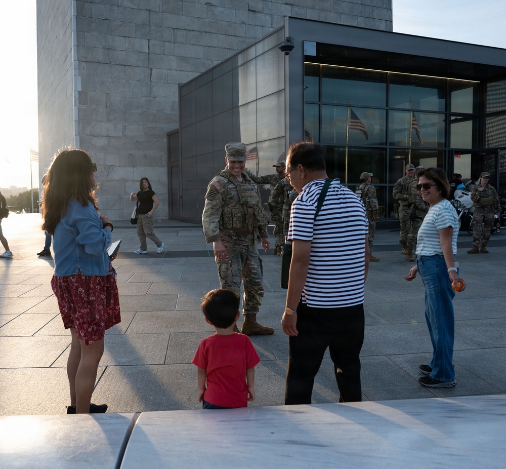 A U.S. Army soldier in full combat uniform and gear is interacting with a civilian family, including a young child, near what appears to be the base of the Washington Monument in Washington, D.C. The soldier is smiling and engaging warmly with the group. Additional soldiers in camouflage uniforms are visible in the background near a glass-fronted building entrance.