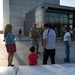A U.S. Army soldier in full combat uniform and gear is interacting with a civilian family, including a young child, near what appears to be the base of the Washington Monument in Washington, D.C. The soldier is smiling and engaging warmly with the group. Additional soldiers in camouflage uniforms are visible in the background near a glass-fronted building entrance.