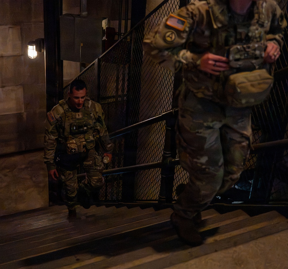Two military personnel in US Army MultiCam/OCP camouflage uniforms are ascending a staircase in a dimly lit environment. Both soldiers are wearing tactical gear including plate carriers and equipment pouches. The foreground soldier displays a US flag patch and what appears to be a First Army patch on their shoulder.