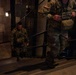Two military personnel in US Army MultiCam/OCP camouflage uniforms are ascending a staircase in a dimly lit environment. Both soldiers are wearing tactical gear including plate carriers and equipment pouches. The foreground soldier displays a US flag patch and what appears to be a First Army patch on their shoulder.
