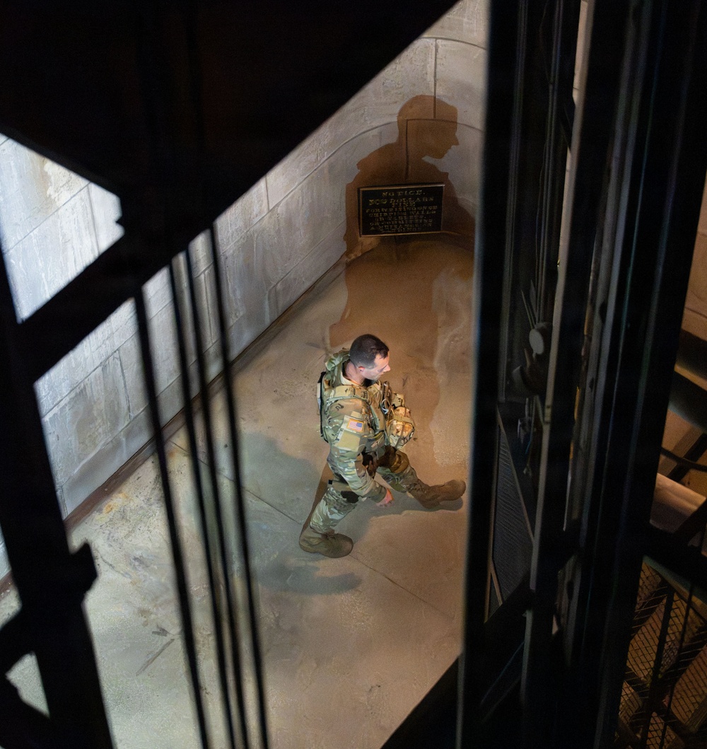 A U.S. military soldier in full camouflage uniform and tactical gear is photographed from an elevated angle, walking through what appears to be a stone corridor or hallway of a large institutional or government building. The soldier's shadow is cast dramatically on the stone wall behind them, and a bronze or brass notice placard is visible on the wall. The image is taken through metal railings, suggesting the photographer is on a staircase or elevated platform above.