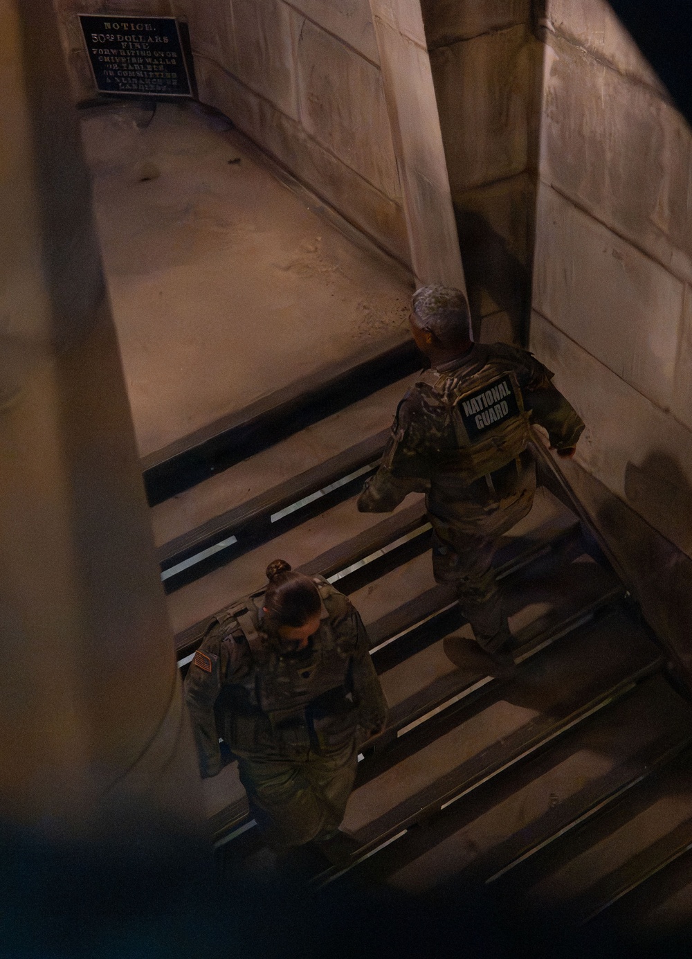 Two National Guard personnel in full tactical gear and camouflage uniforms are ascending a stone staircase, viewed from above at night or in low-light conditions. A historical notice sign is visible on the stone wall to the left, partially legible, warning of a fine. The setting appears to be a historic government or public building with large stone masonry walls.