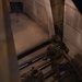 Two National Guard personnel in full tactical gear and camouflage uniforms are ascending a stone staircase, viewed from above at night or in low-light conditions. A historical notice sign is visible on the stone wall to the left, partially legible, warning of a fine. The setting appears to be a historic government or public building with large stone masonry walls.