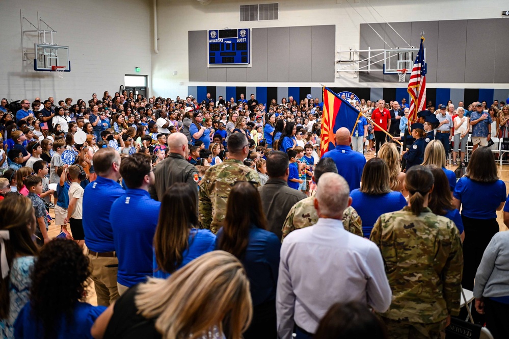A large patriotic assembly is taking place inside a school gymnasium, with hundreds of students, parents, and military personnel gathered together. The scoreboard identifies the home team as the 'Aviators,' and flags including the American flag and the Arizona state flag are prominently displayed. Military personnel in camouflage uniforms and civilians in blue shirts are present, suggesting a Veterans Day or military appreciation event.